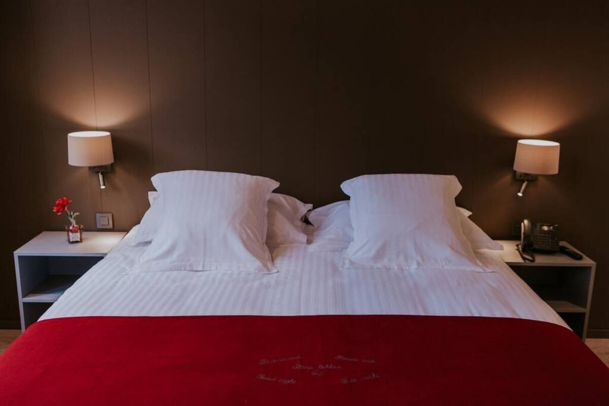 Una cama bien tendida con sábanas blancas y una manta roja, flanqueada por dos mesitas de noche con lámparas y un pequeño jarrón de flores.