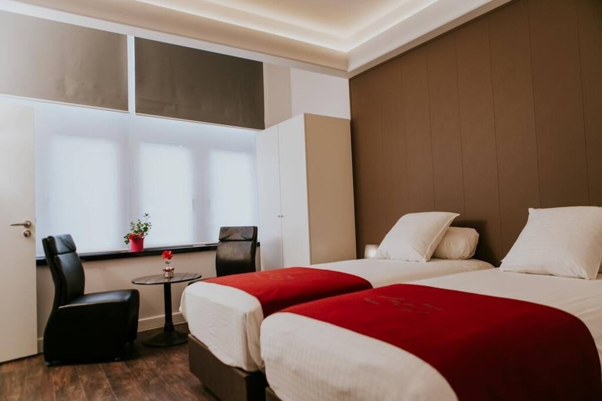 Habitación de hotel con dos camas, una mesita con una flor roja, una silla y una ventana con persianas.