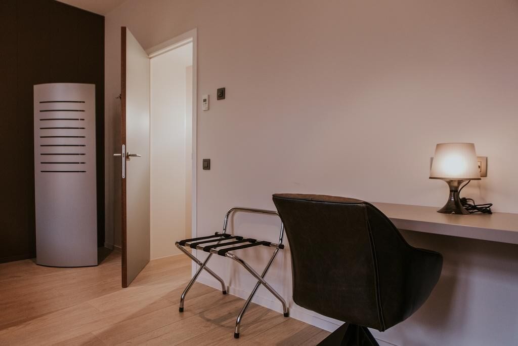 Een moderne kamer met een houten vloer, een zwarte lederen fauteuil, een klein tafeltje met een lamp en een witte deur.