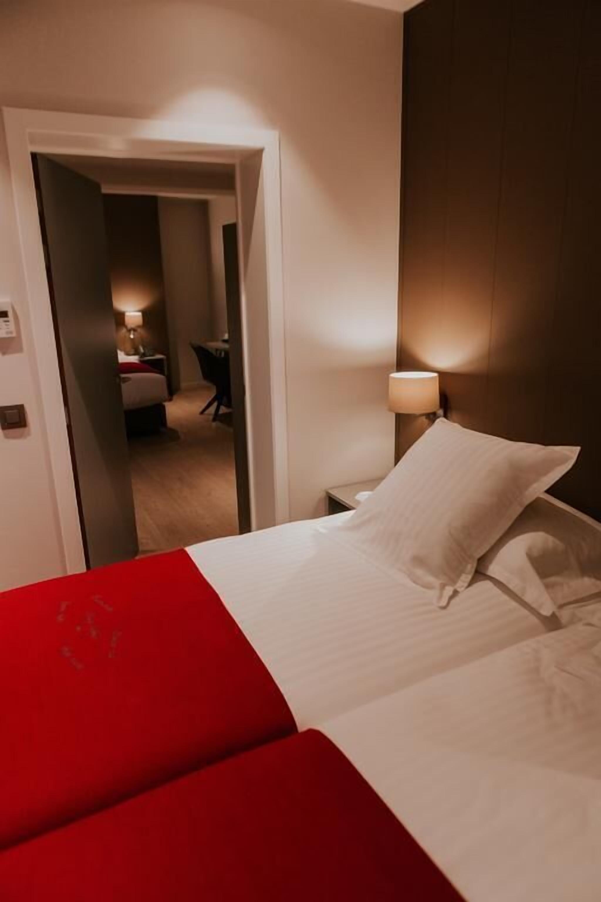 A hotel room with a bed, a nightstand, a lamp, and a door leading to another room.