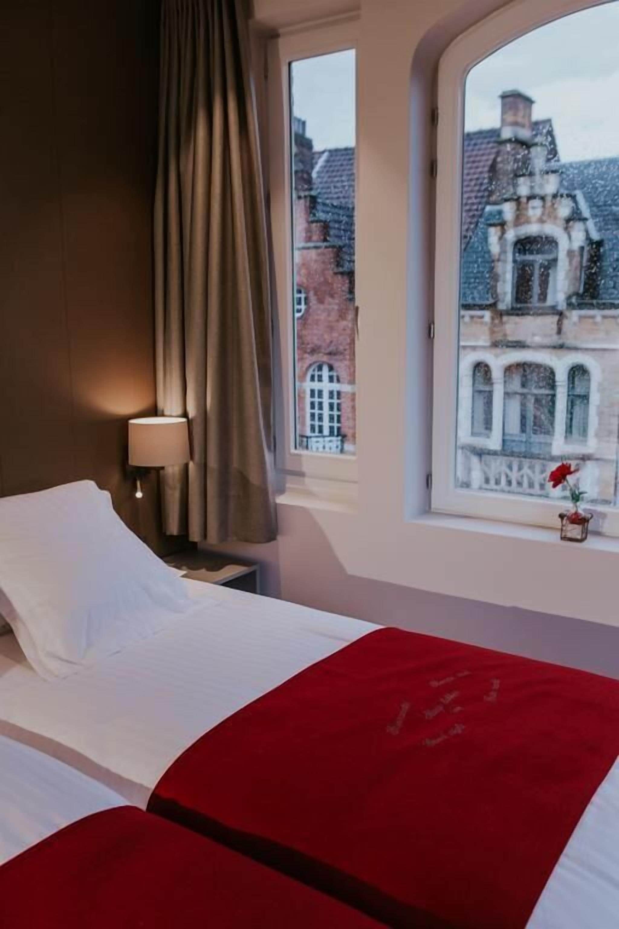 A hotel room with a bed, a nightstand, a lamp, and a window with a view of a building.
