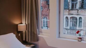 A hotel room with a bed, a nightstand, a lamp, and a window with a view of a building.