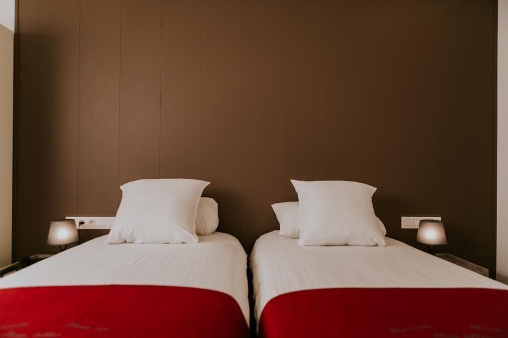 Dos camas individuales con sábanas blancas y cubrecamas rojos, ubicadas frente a una pared marrón con diseño de paneles de madera.