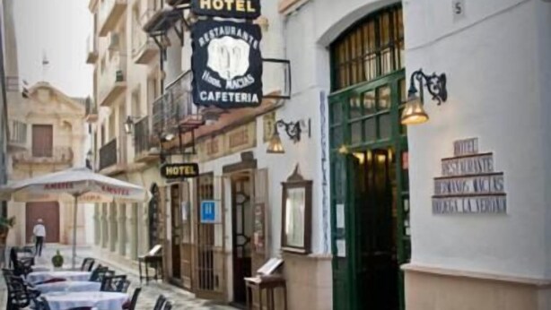 An outdoor cafe area with tables and chairs, a hotel sign, and a building with a green door. - Hermanos Macías Hotel (Ronda)