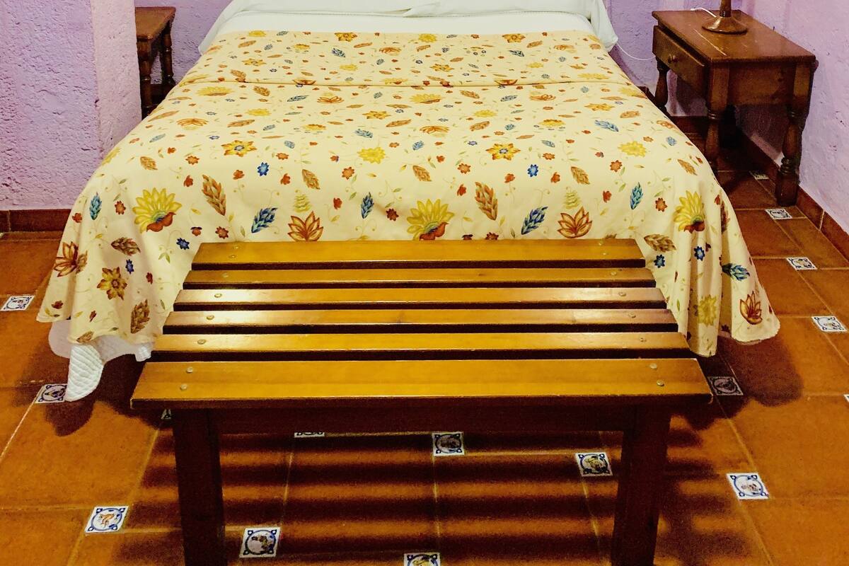 A bedroom with a wooden bed, floral bedspread, two bedside lamps, a nightstand, and a bench.