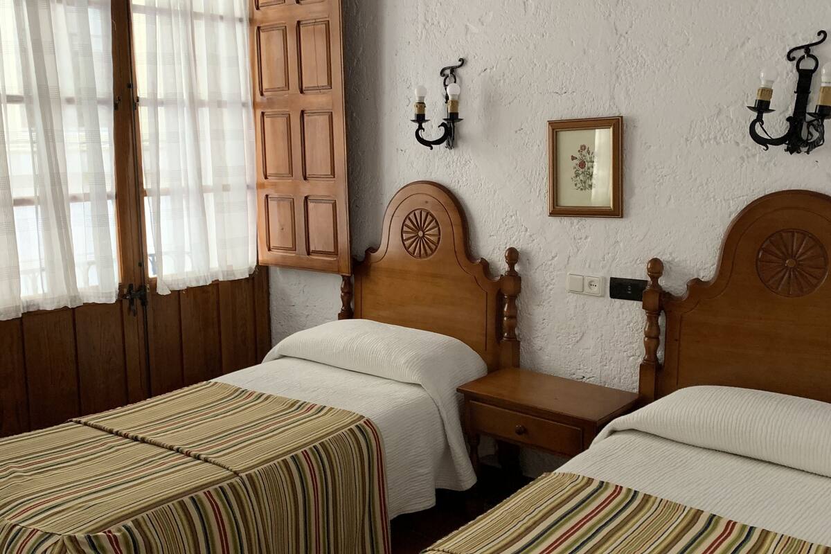 A room with two wooden beds, a wooden wardrobe, a small table, and a wall-mounted light fixture.