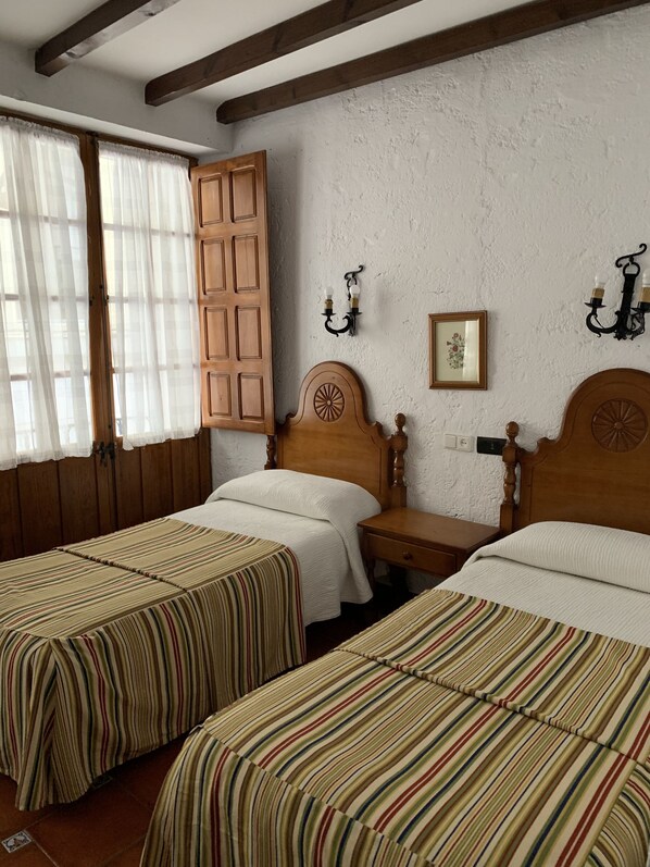 A room with two single beds, wooden headboards, a nightstand, a wall-mounted light fixture, and a window with curtains.