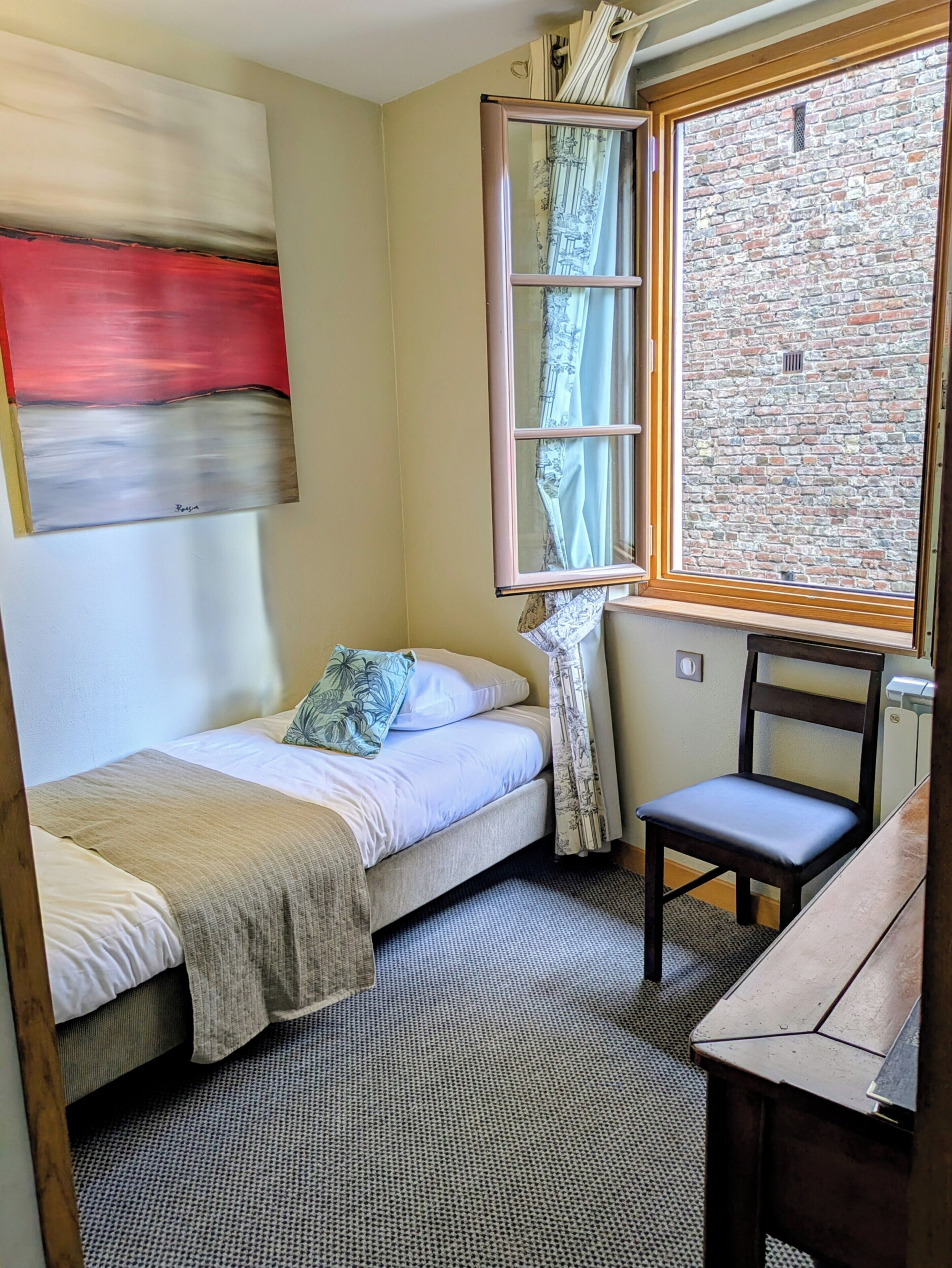 A small room with a bed, a chair, a window with curtains, and a brick wall.