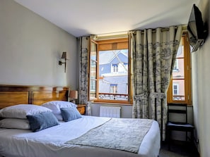 A hotel room with a bed, a nightstand, a lamp, a window with curtains, and a TV mounted on the wall. - Hôtel Entre Terre et Mer (Honfleur)
