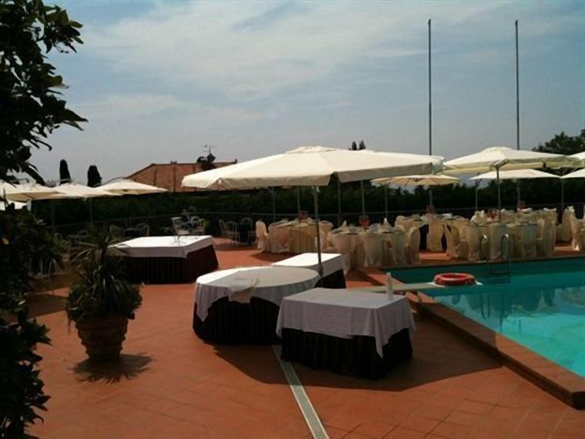 Zona de piscina exterior con mesas redondas y sombrillas, rodeada de plantas en macetas y un cielo despejado.