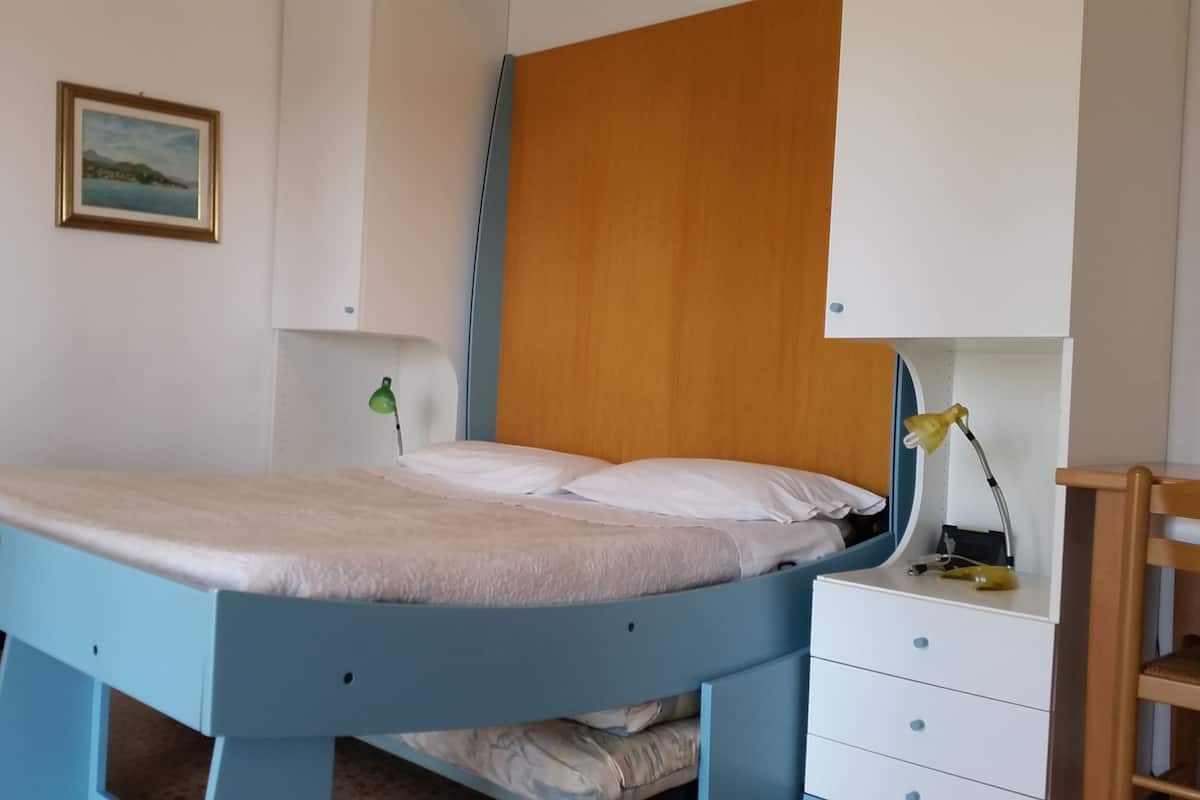 A bedroom with a blue bed frame, white bedding, a wooden headboard, a bedside table with a lamp, and a small wooden chair.