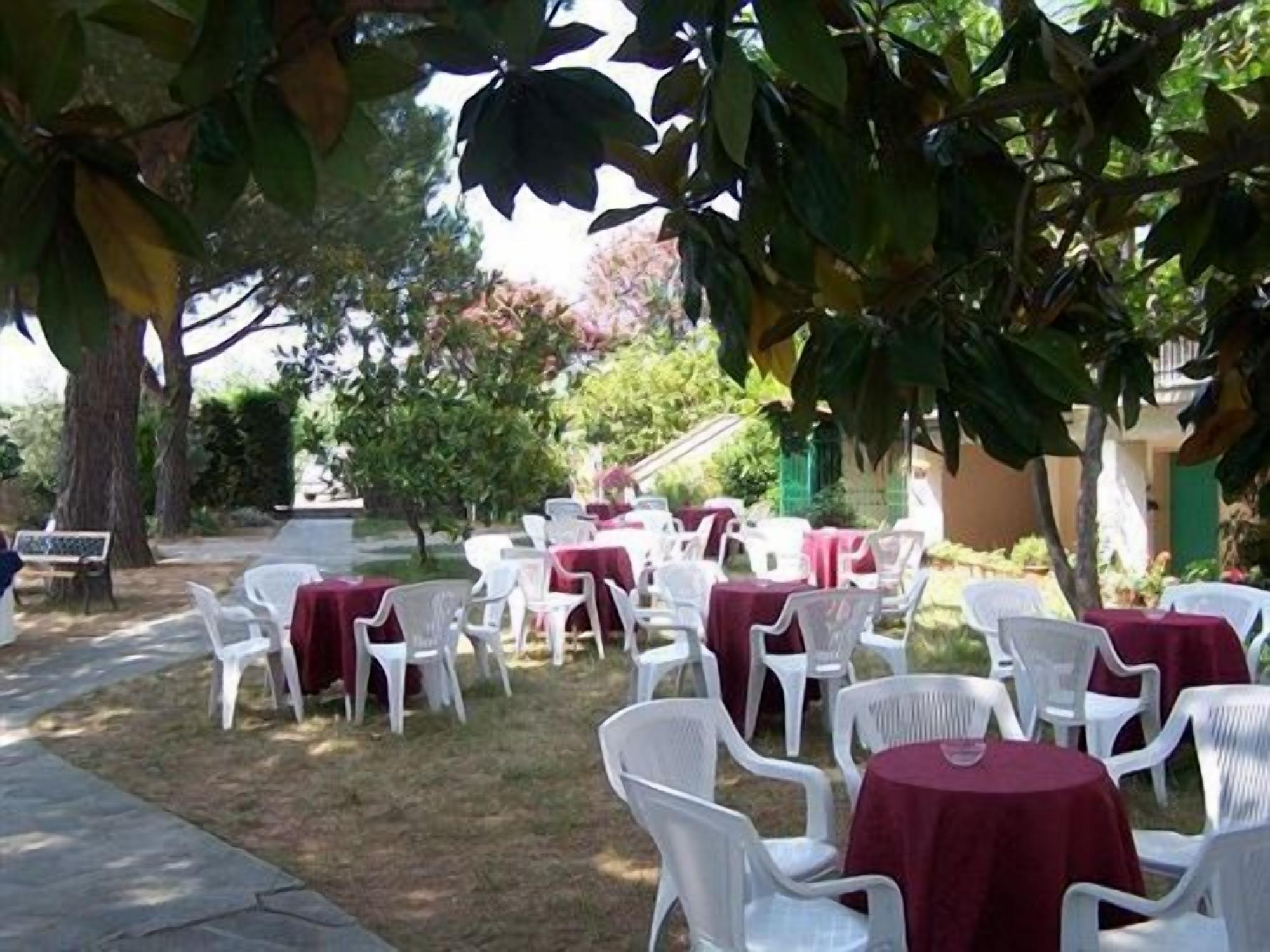 An outdoor seating area with white plastic chairs and tables covered with red tablecloths, surrounded by greenery and trees.