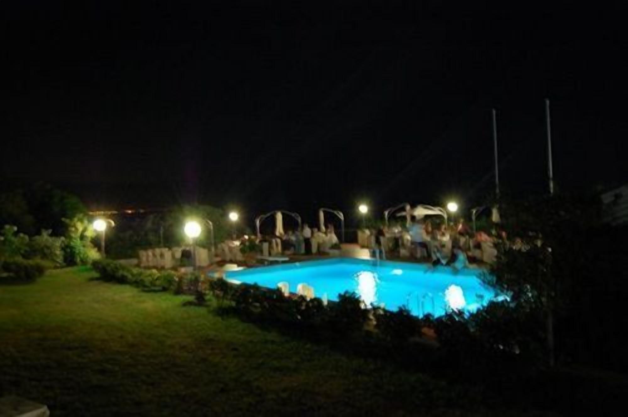 Zona de piscina nocturna con una piscina cristalina, rodeada de césped e iluminada por luces colgantes.