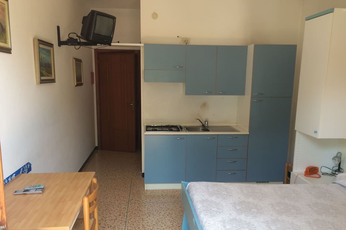 A compact room with a bed, a small table, a kitchenette, and a TV mounted on the wall.