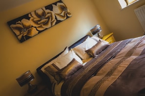 A bed with a striped blanket and multiple pillows, a nightstand with a lamp, and a wall-mounted painting of flowers. - The Lindley Suite - Simple2let Serviced Apartments (Huddersfield)