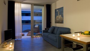 A modern living room with a sofa, a dining table with candles, and a view through a glass door to an outdoor balcony. - Residence Hotel Hungaria (Grado)
