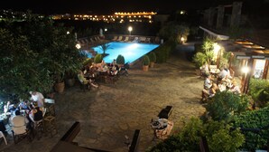 An outdoor pool area with stone flooring, surrounded by potted plants and tables with chairs. - Golden Apartments (Agios Nikolaos)