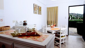 A kitchen with white cabinetry, a dining table with chairs, and a view of a garden through an open door. - Golden Apartments (Agios Nikolaos)