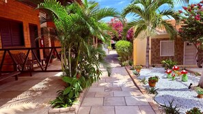 Um caminho leva a um edifício com fachada de tijolos, cercado por vegetação exuberante e plantas decorativas.
