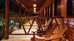 A covered outdoor seating area with wooden chairs and tables, a brick wall, and hanging lights. - Pousada Jardim dos Orixás (Aracati)