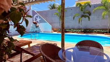 Uma área à beira da piscina com mesa de vidro e cadeiras de vime, parede de pedra com um mural de estrelas e uma piscina azul cristalina.