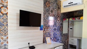 A hotel room with a flat-screen TV mounted on the wall, a small refrigerator, a desk with a power outlet, and a colorful tile backsplash. - Pousada Jardim dos Orixás (Aracati)