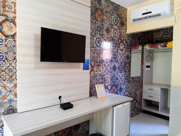 A hotel room with a flat-screen TV mounted on the wall, a small refrigerator, a desk with a power outlet, and a colorful tile backsplash. - Pousada Jardim dos Orixás (Aracati)