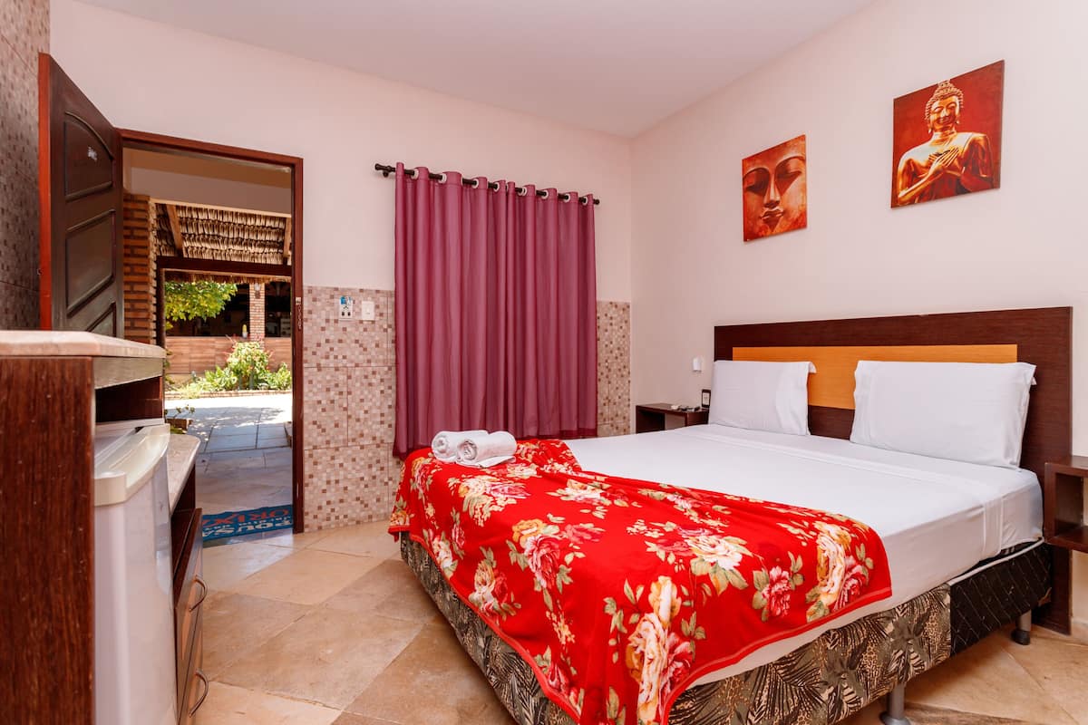 A hotel room with a bed, a wooden headboard, a nightstand, a wall-mounted TV, a window with curtains, and two Buddha paintings on the wall.