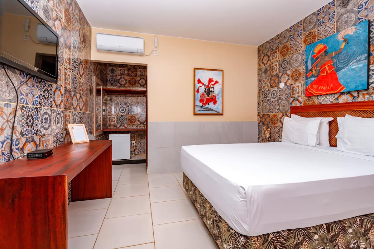 A hotel room with a large bed, a wooden desk, a television, and a wall with decorative tiles and artwork.