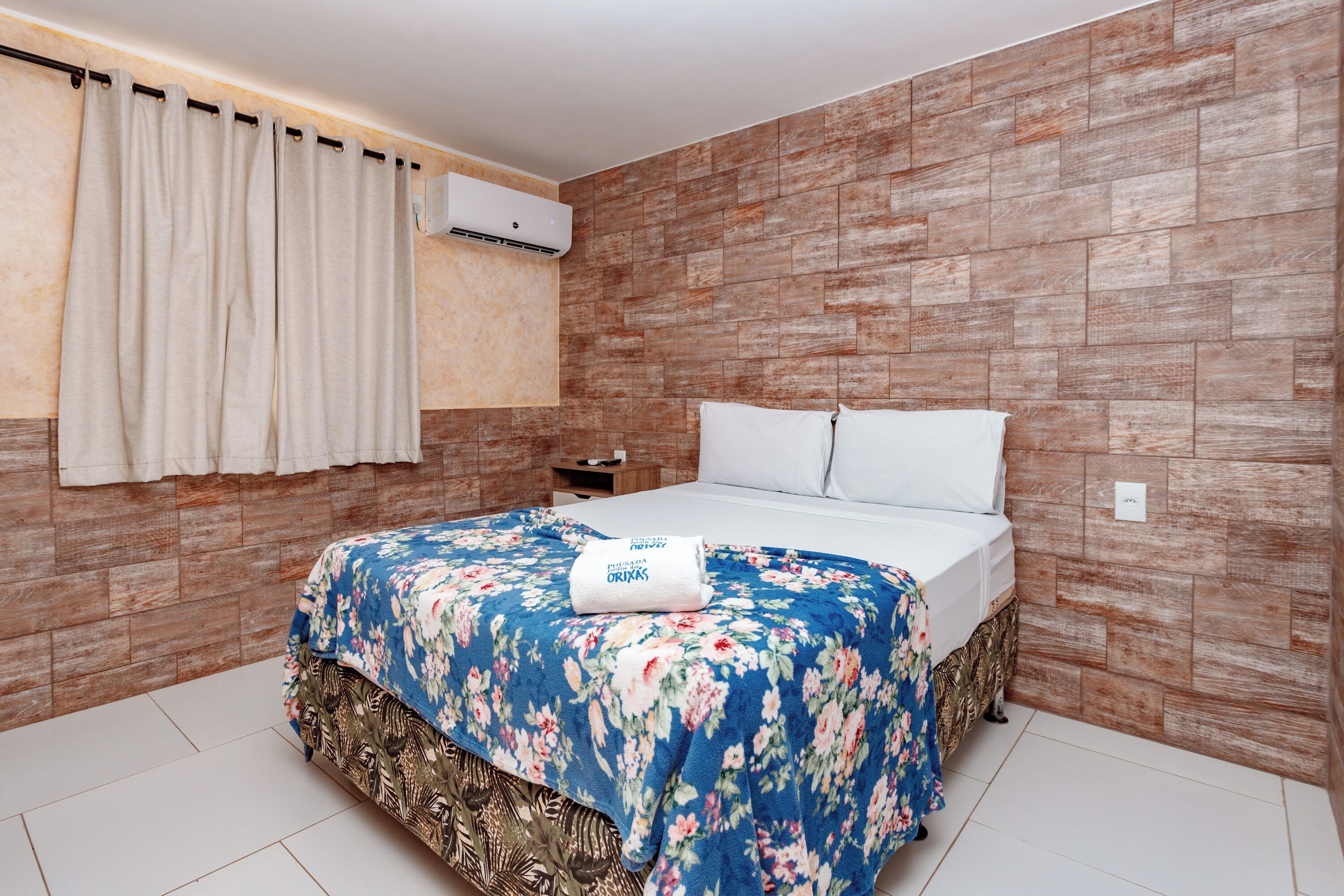 Un dormitorio con cama, mesita de noche, aire acondicionado de pared y ventana con cortinas.
