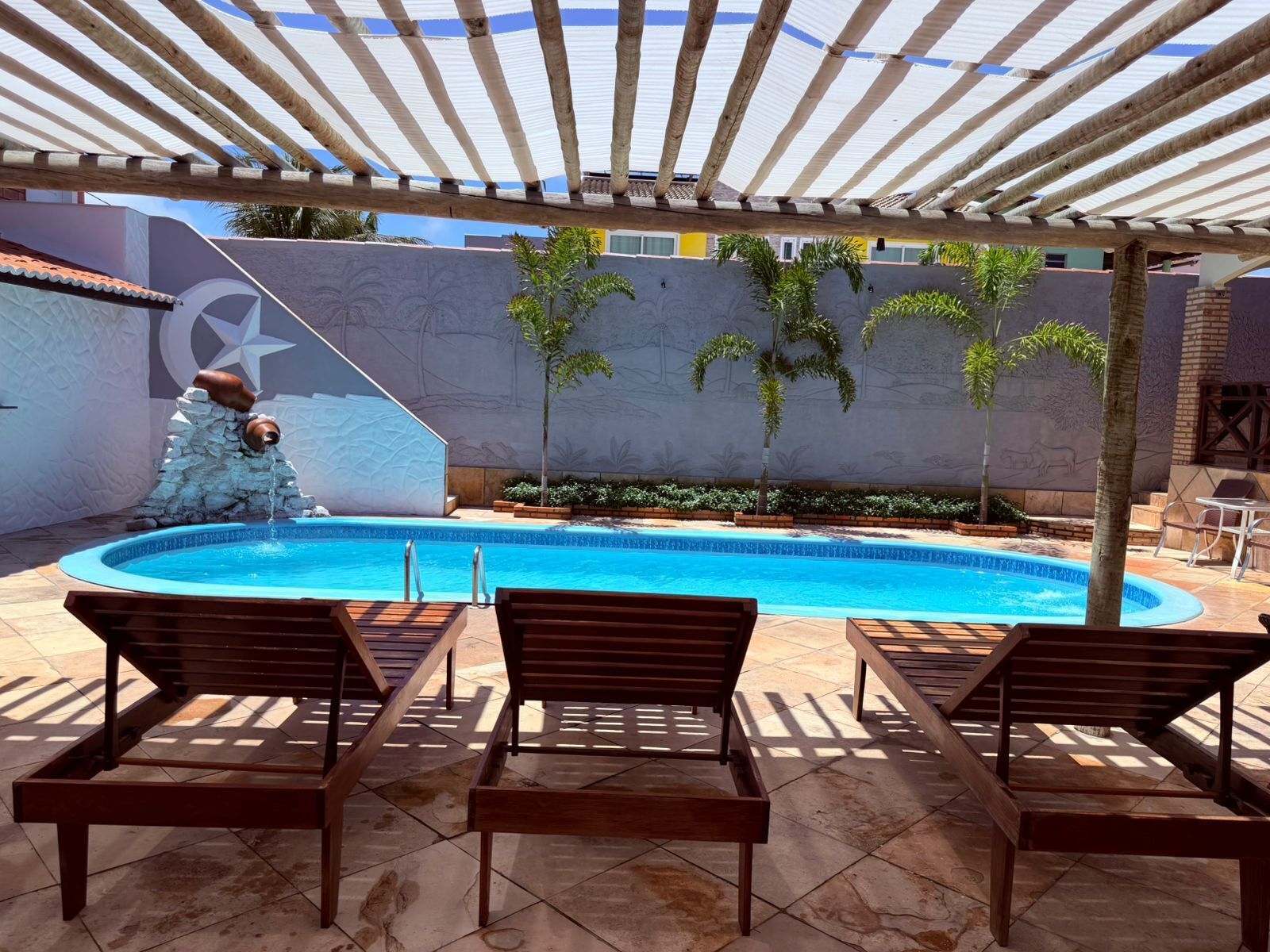 Piscina com uma fonte de pedra, rodeada por espreguiçadeiras de madeira e uma área coberta com toldo listrado.
