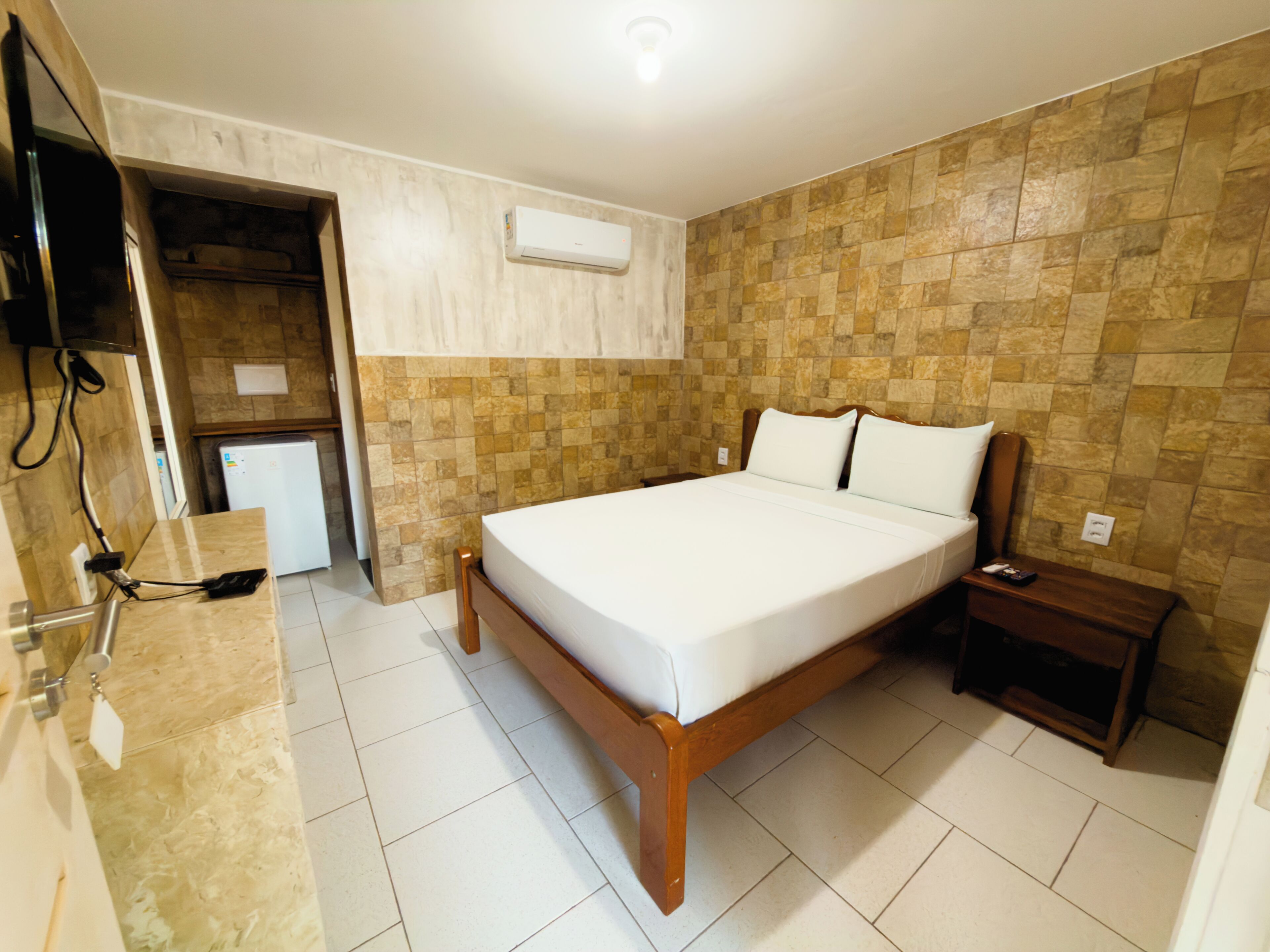 A hotel room with a bed, a bedside table, a TV, and a built-in air conditioning unit.