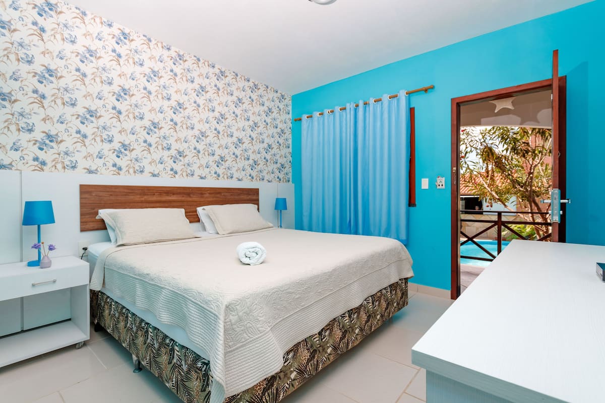 A bedroom with a bed, nightstands, a desk, a window with curtains, and a view of a pool.