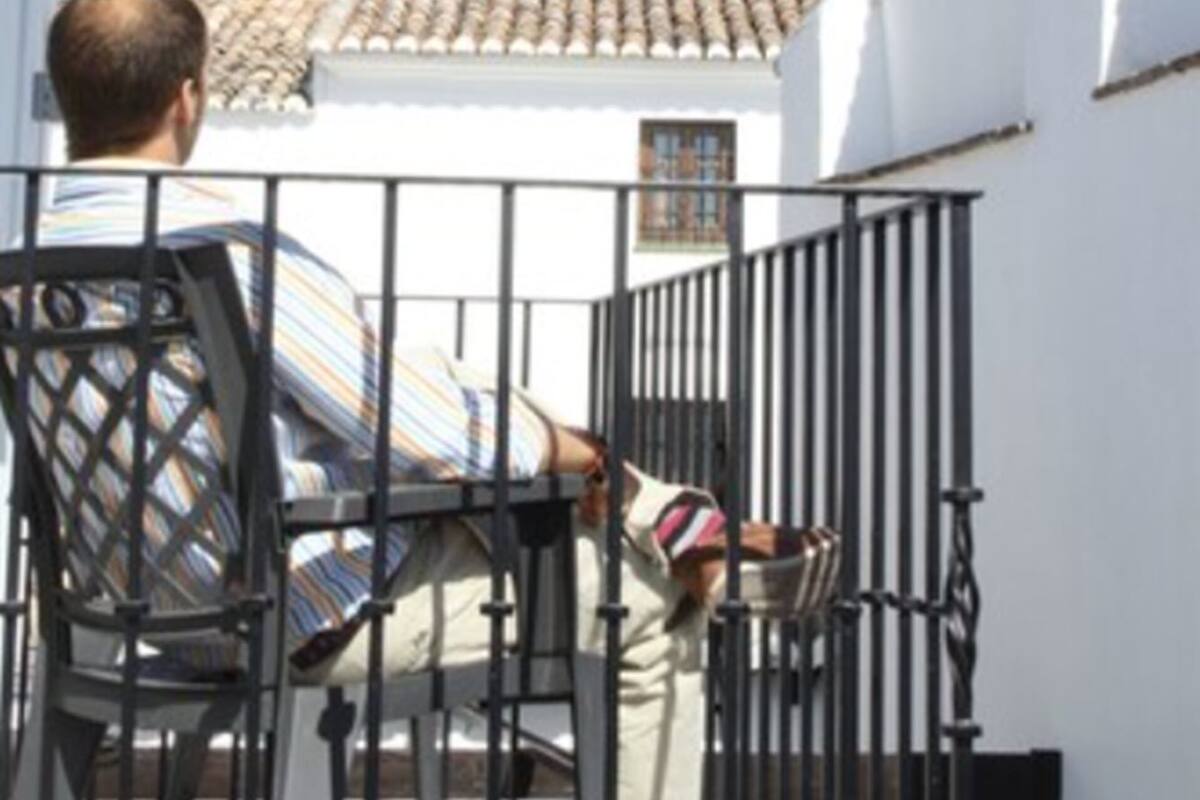 A man sitting at a table with a striped cushion, a metal railing, and a building with a tiled roof in the background.