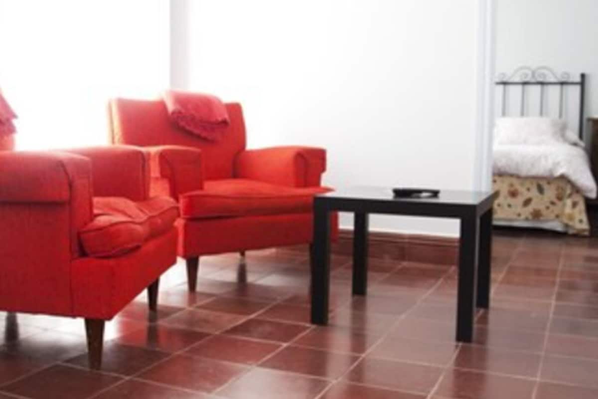 A room with two red armchairs, a small black table, and a bed with a white headboard.