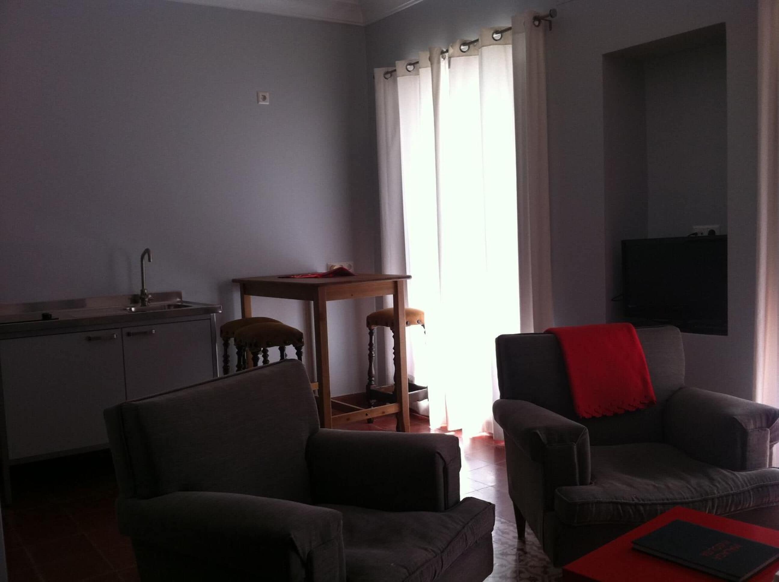 A living room with two armchairs, a small table, a TV mounted on the wall, and a kitchen area with a sink and cabinets.