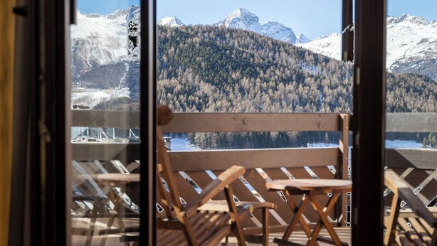 A balcony with wooden chairs and a table, overlooking a snowy mountain and forested area. - Hotel Rosatsch - Stammhaus (Pontresina)