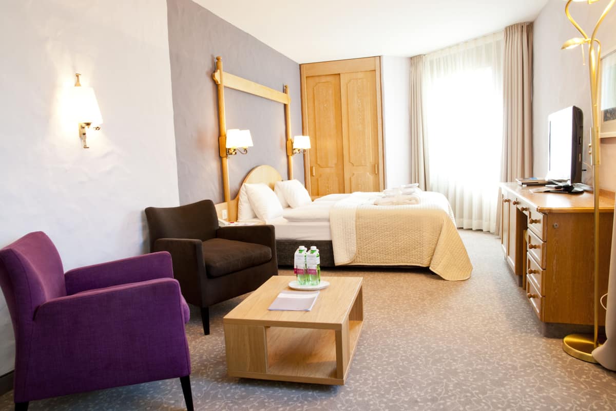 Quarto de hotel com cama, escrivaninha, cadeira, mesa de centro e televisão.