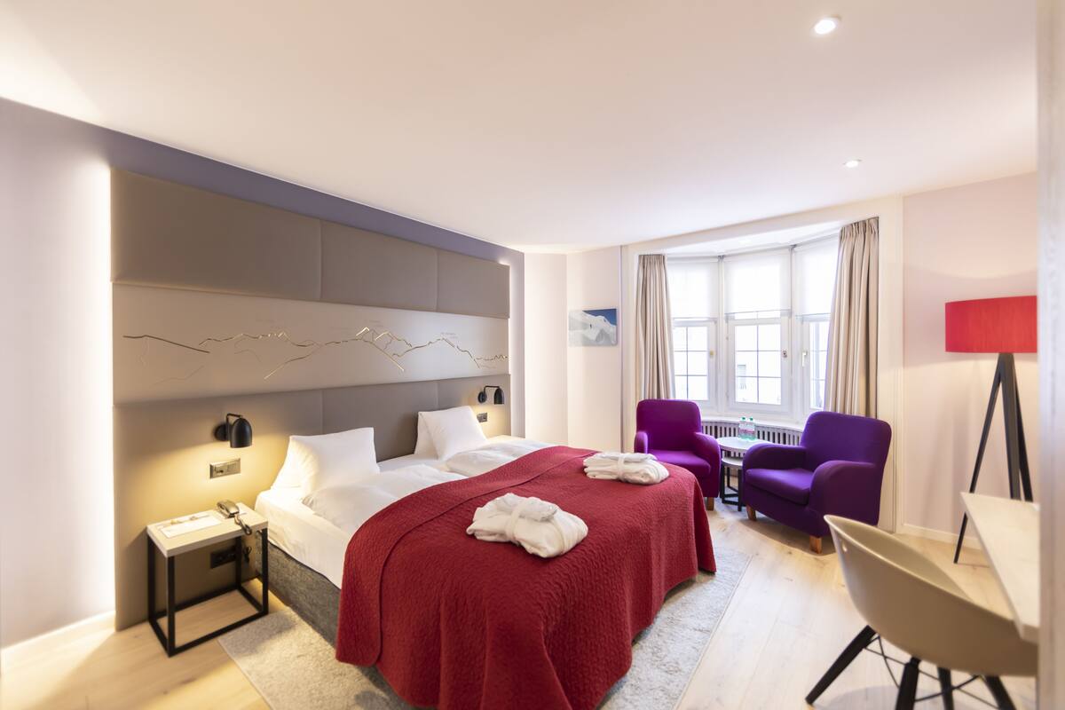 Quarto de hotel moderno com uma cama grande, uma escrivaninha, uma cadeira e uma janela com cortinas.
