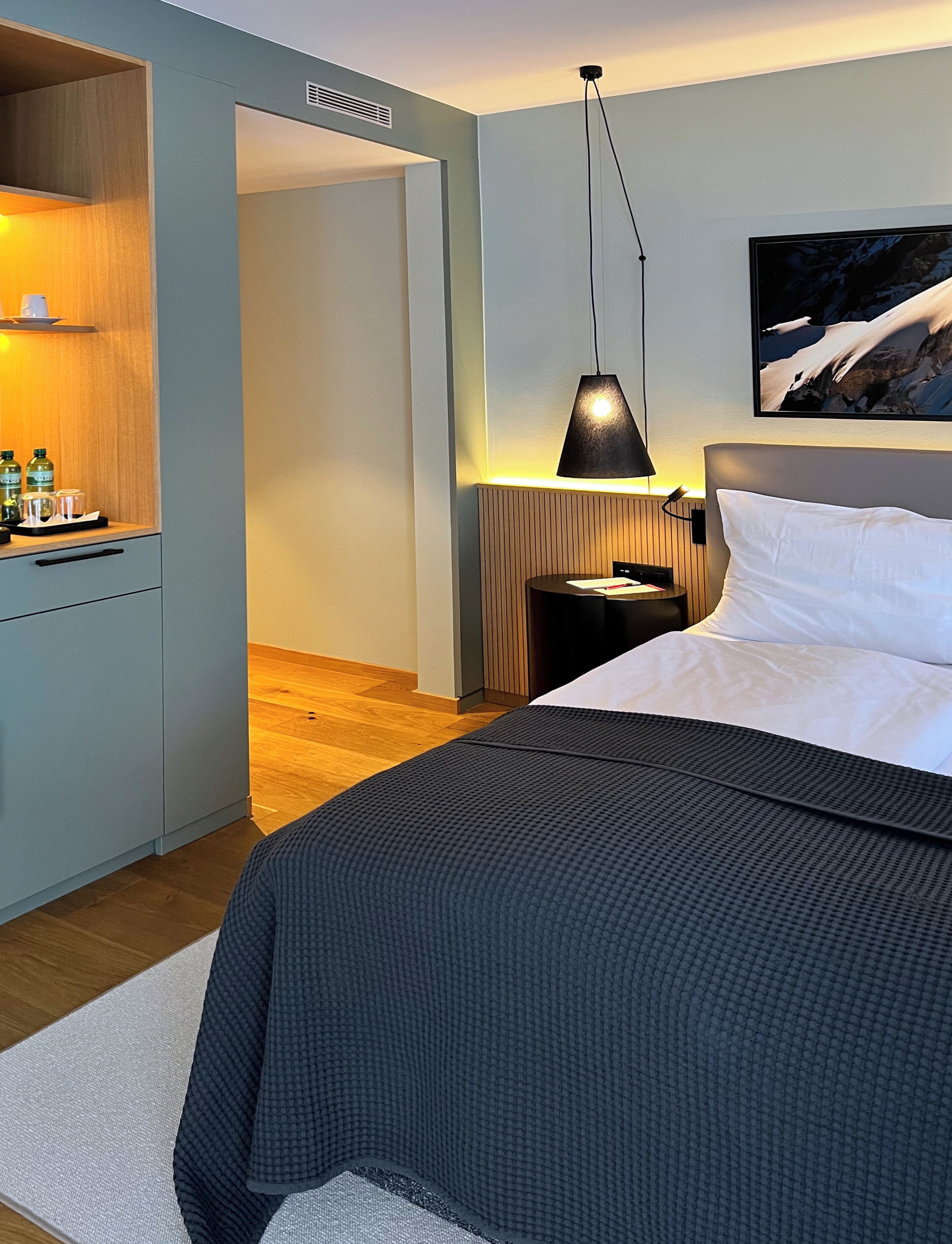 A modern hotel room with a large bed, a bedside table, a desk with a chair, a lamp, and a painting on the wall.