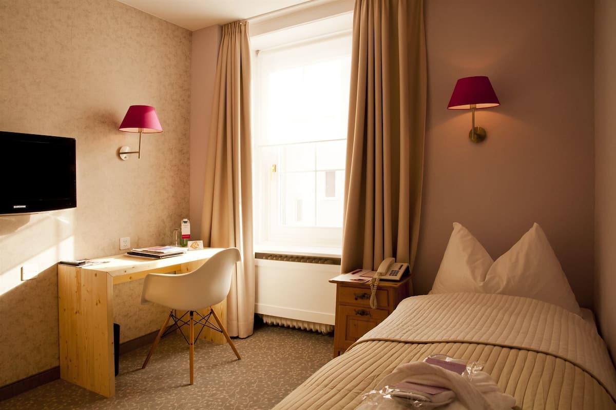 Quarto de hotel com cama, escrivaninha, cadeira, televisão e duas luminárias.