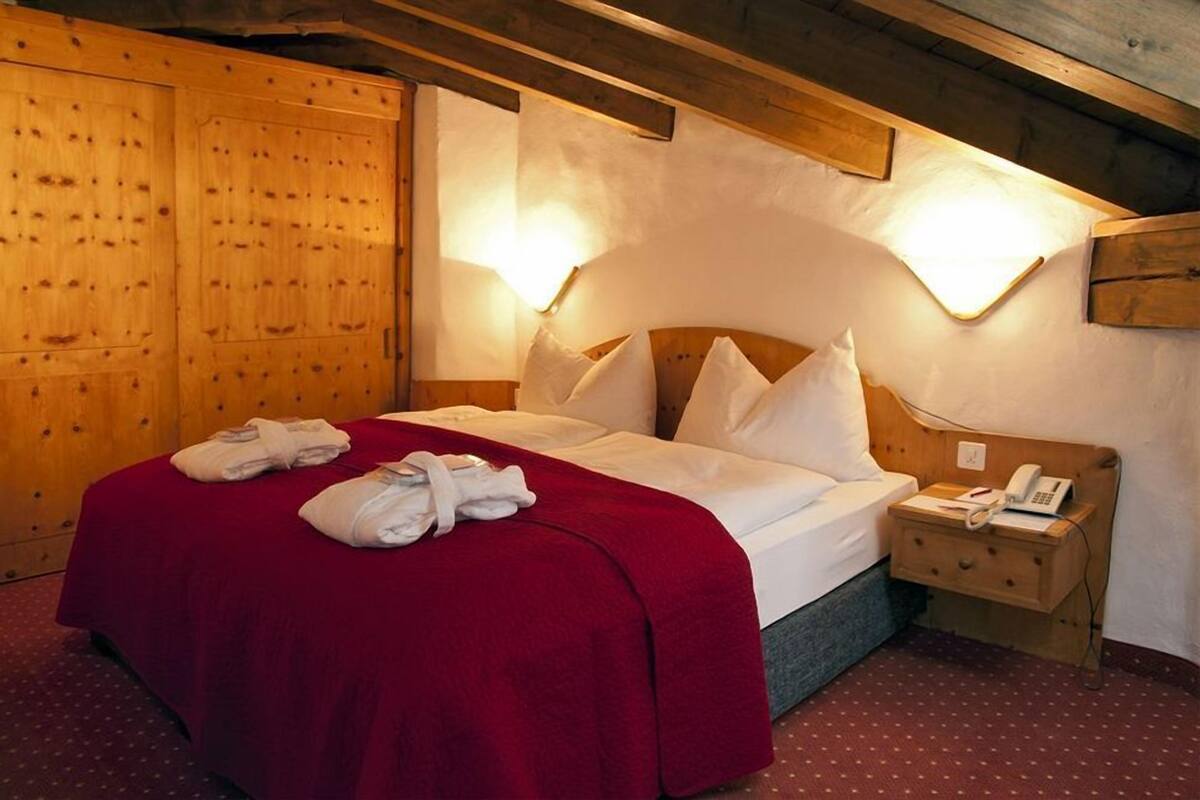 Quarto de hotel com cama de madeira, manta vermelha, mesa de cabeceira com telefone e guarda-roupa de madeira.