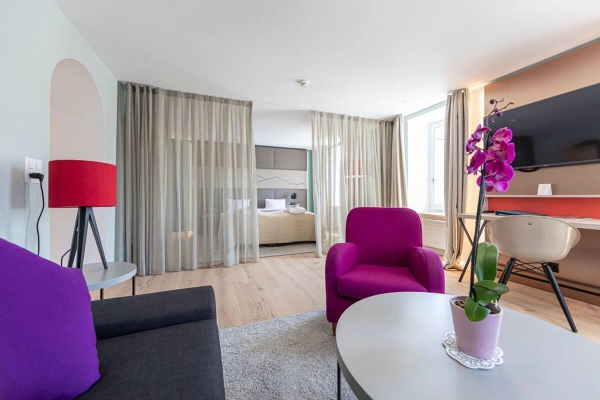 Quarto de hotel moderno com cama, escrivaninha, televisão, poltrona roxa, mesinha e um vaso com planta.