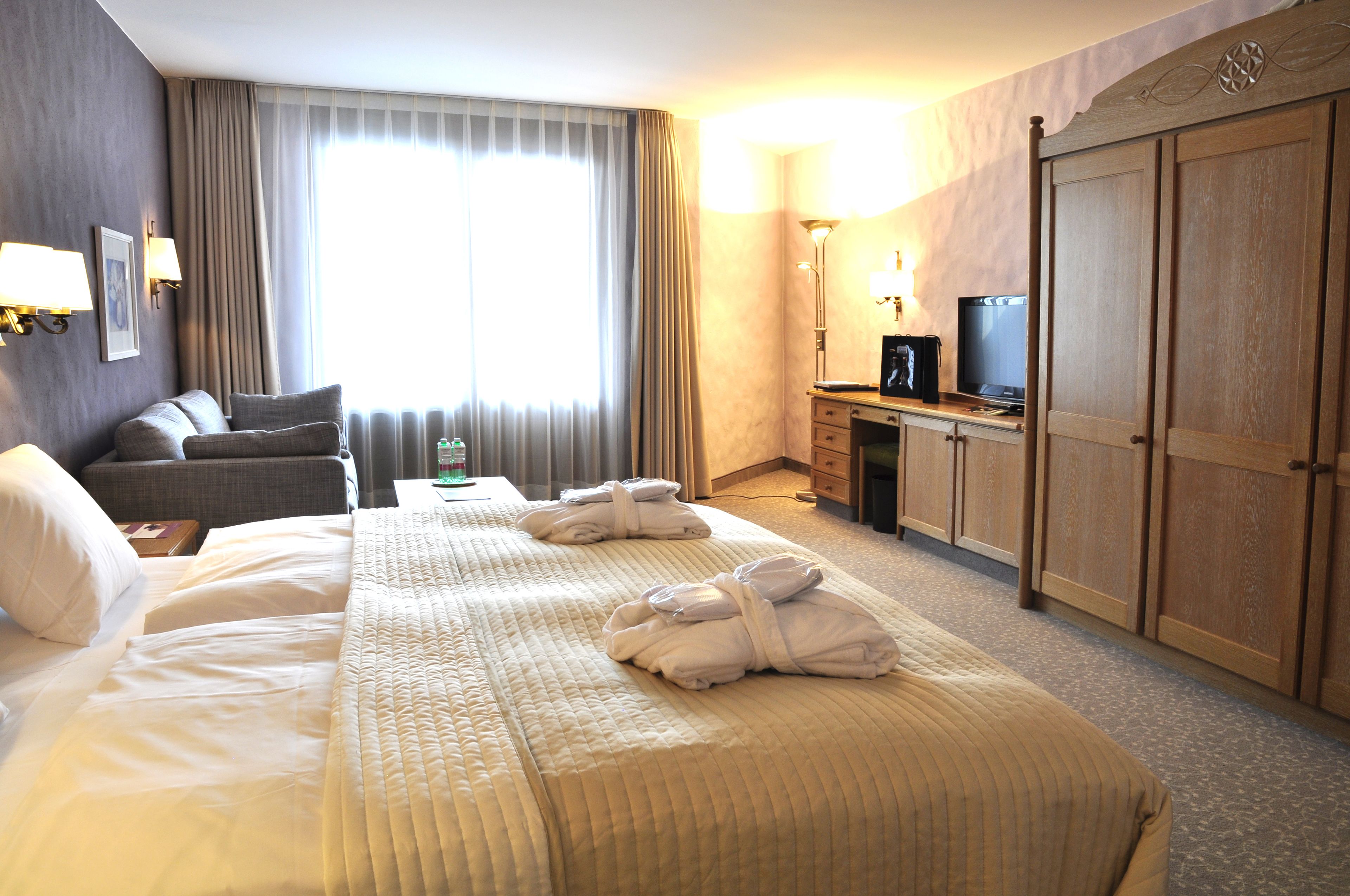 A hotel room with a large bed, two armchairs, a desk with a TV, and a wardrobe.