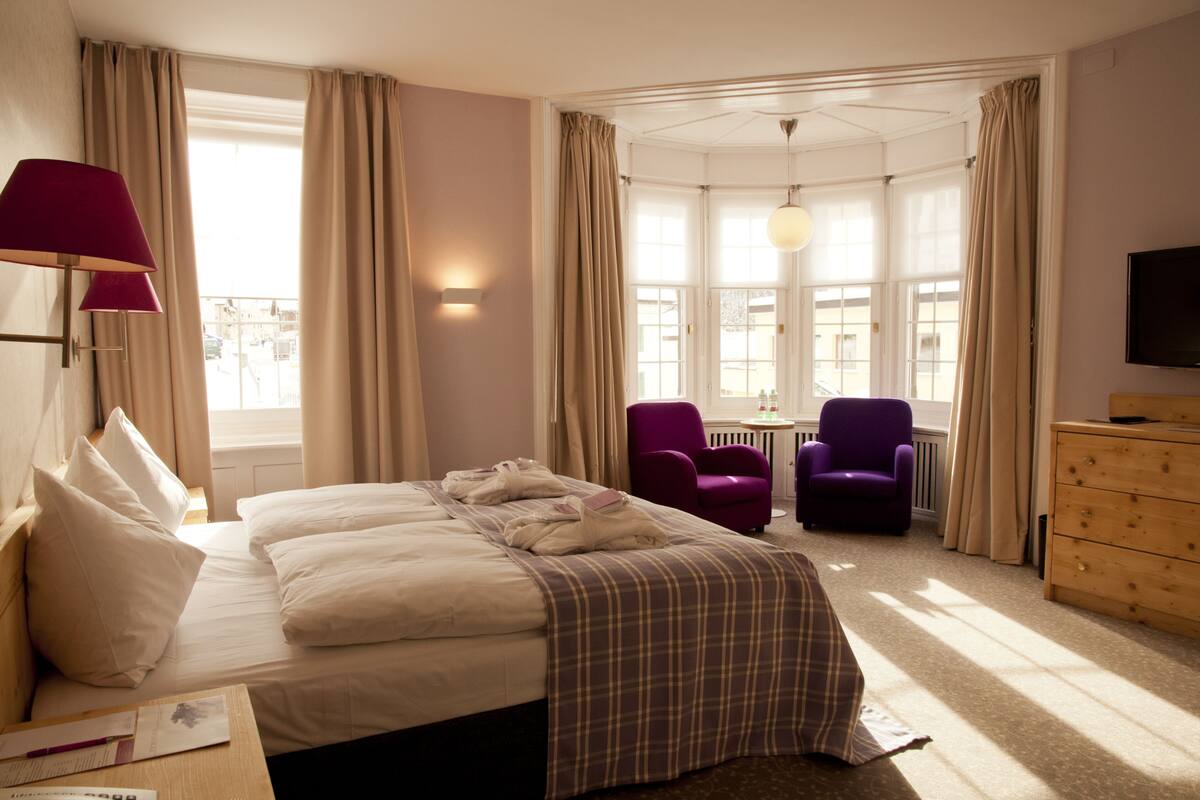 Quarto de hotel com cama, duas poltronas, cómoda de madeira, televisão e janela com cortinas.