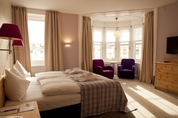 Habitación de hotel con cama, dos sillones, cómoda de madera, televisor y ventana con cortinas.