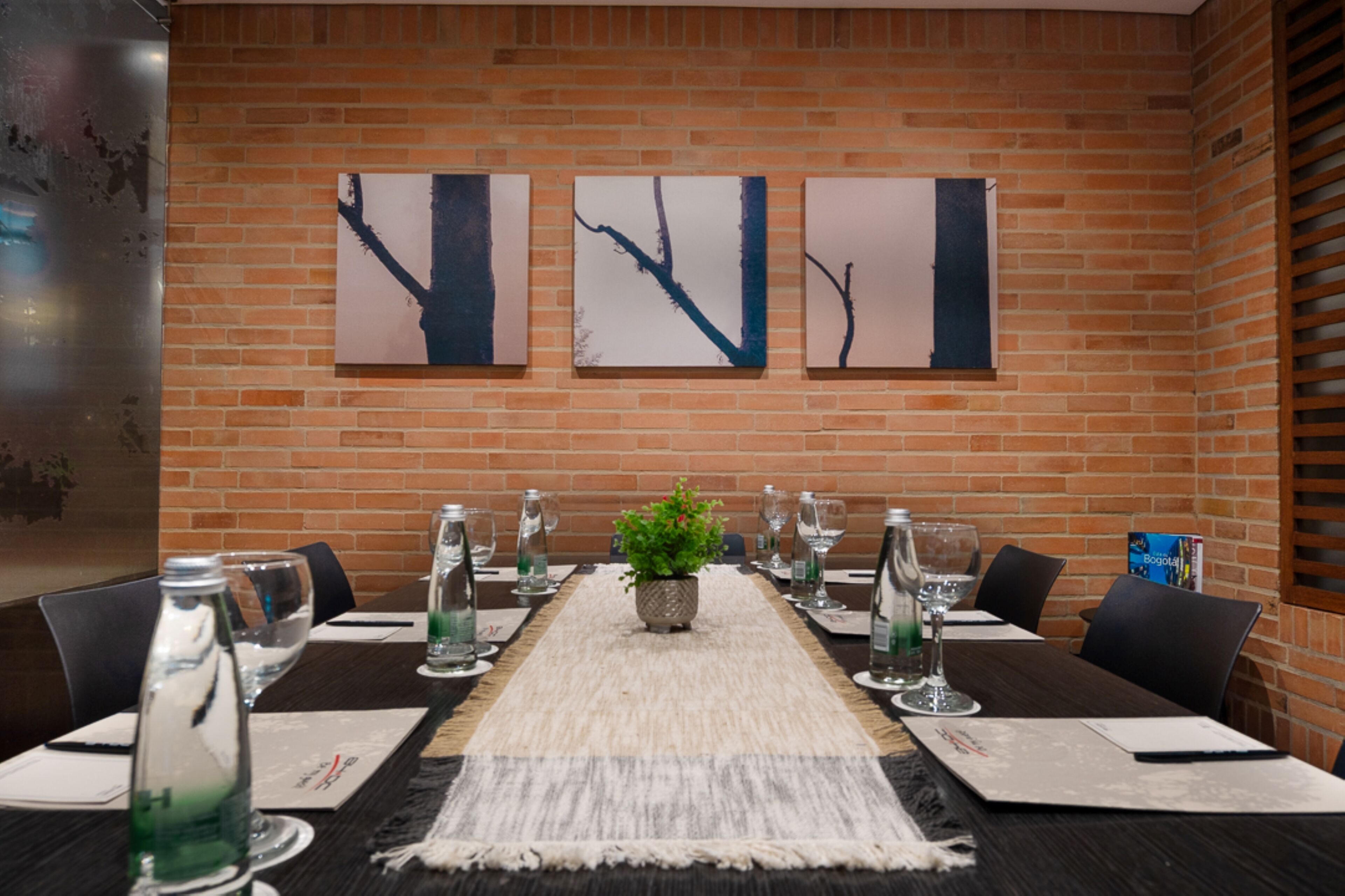 A conference room with a long table set for a meeting, a potted plant, and three abstract paintings on the brick wall.