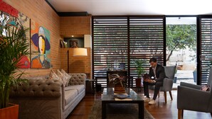 A modern living room with a sofa, coffee table, and chairs, featuring a large window with blinds and a view of the outdoors. - 84 DC Bogotá By OxoHotel (Bogotá)