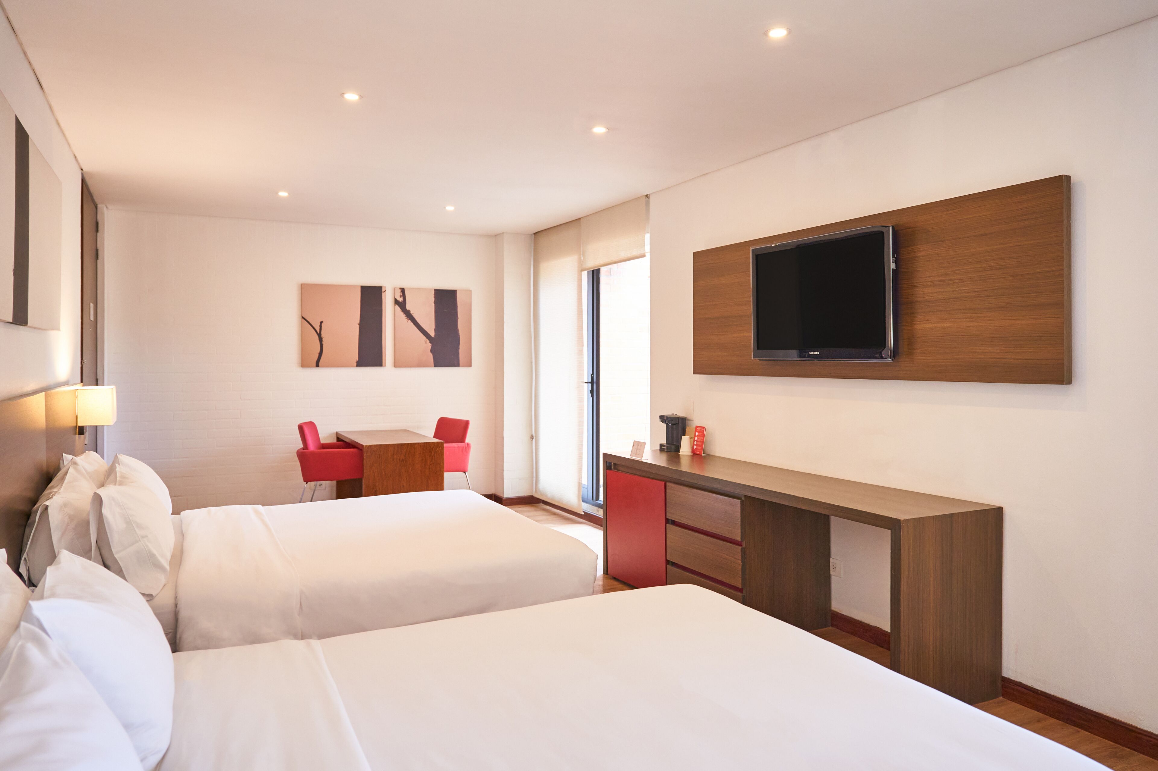 Habitación de hotel con dos camas, un escritorio, un televisor montado en la pared y una silla roja.