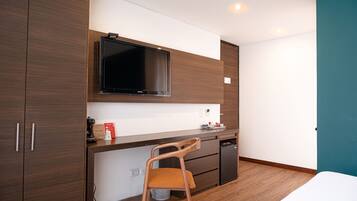 A compact room with a wooden desk, a television mounted above it, a small kitchenette, and a single bed.